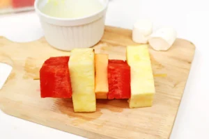 Fruit skewers with watermelon and pineapple on a wooden board, with a white bowl in the background.