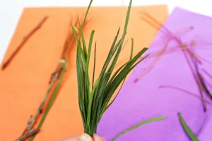 Close-up of a hand holding a small bundle of green grass against an orange and purple background, foreground focus on the needles.