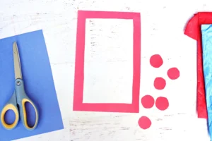 Scissors on a blue sheet of paper next to a red paper frame and scattered red dots on a white wooden surface, with red tissue nearby—craft supplies on a desk.