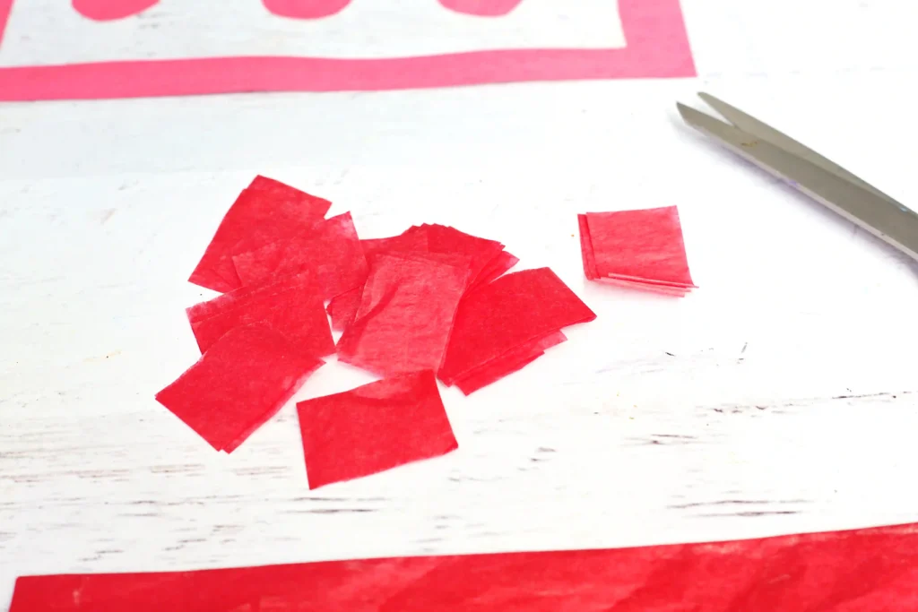Scattered red tissue paper squares with a pair of scissors on a white wooden surface, suggesting a craft project.