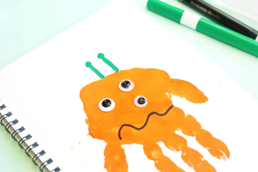 Letter M Handprint Craft - Cultivating Brilliant Minds Orange handprint monster with googly eyes on a white notebook page for this handprint art