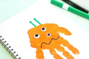 Orange handprint monster with googly eyes on a white notebook page for this handprint art