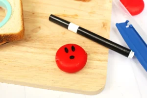 Red round babybel cheese snack with a small black facial feature sits on a light wooden surface beside a black marker for these creative school lunch box ideas