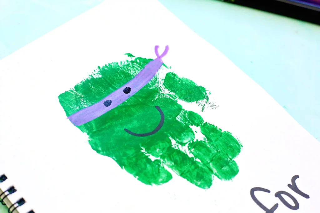 Close-up of a green handprint stamp on white paper with a purple curved line and two black dots to look like a mask.