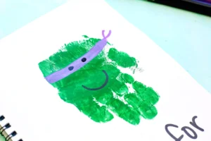 Close-up of a green handprint stamp on white paper with a purple curved line and two black dots to look like a mask.