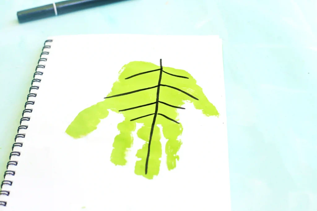 Letter L Leaf Handprint Craft - Cultivating Brilliant Minds Notebook page with a black abstract tree/leaf drawing over a lime-green handprint smear; a pen rests above.