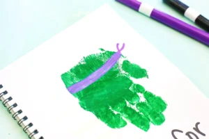 Close-up of a notebook page with a green paint stamp and a purple ribbon/line, and purple and black pens overhead on a light surface.