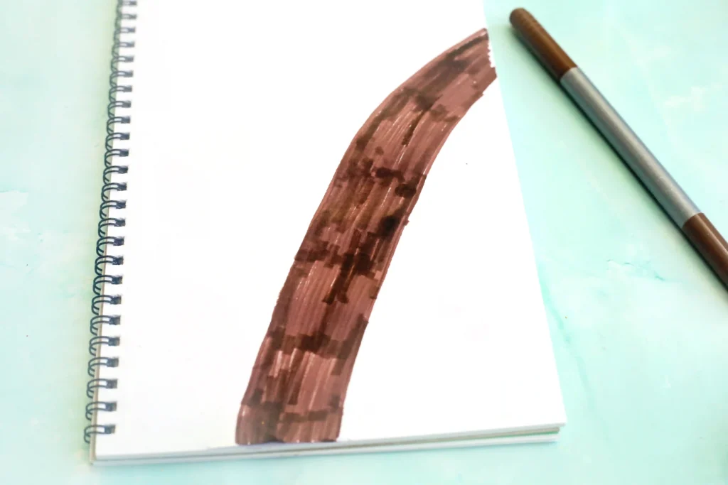 Spiral notebook open with a brown brush stroke across a white page on a light blue surface, pencil nearby on the right for this alphabet craft for kids