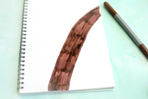 Spiral notebook open with a brown brush stroke across a white page on a light blue surface, pencil nearby on the right for this alphabet craft for kids
