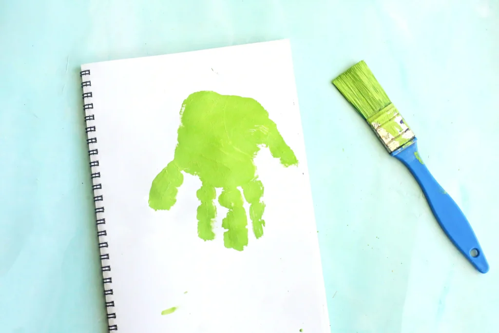 Letter L Leaf Handprint Craft - Cultivating Brilliant Minds White spiral notebook with a lime-green handprint on the page and a blue-green paintbrush on a pale blue surface for this leaf craft for kids
