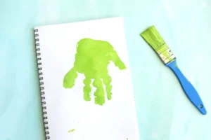 White spiral notebook with a lime-green handprint on the page and a blue-green paintbrush on a pale blue surface for this leaf craft for kids