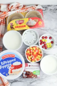 ingredients for the mini turkey pies: Assorted dessert ingredients on a marble counter: graham crackers, whipped topping, candy corn, white nonpareils, and red/yellow candies with an orange striped towel nearby.