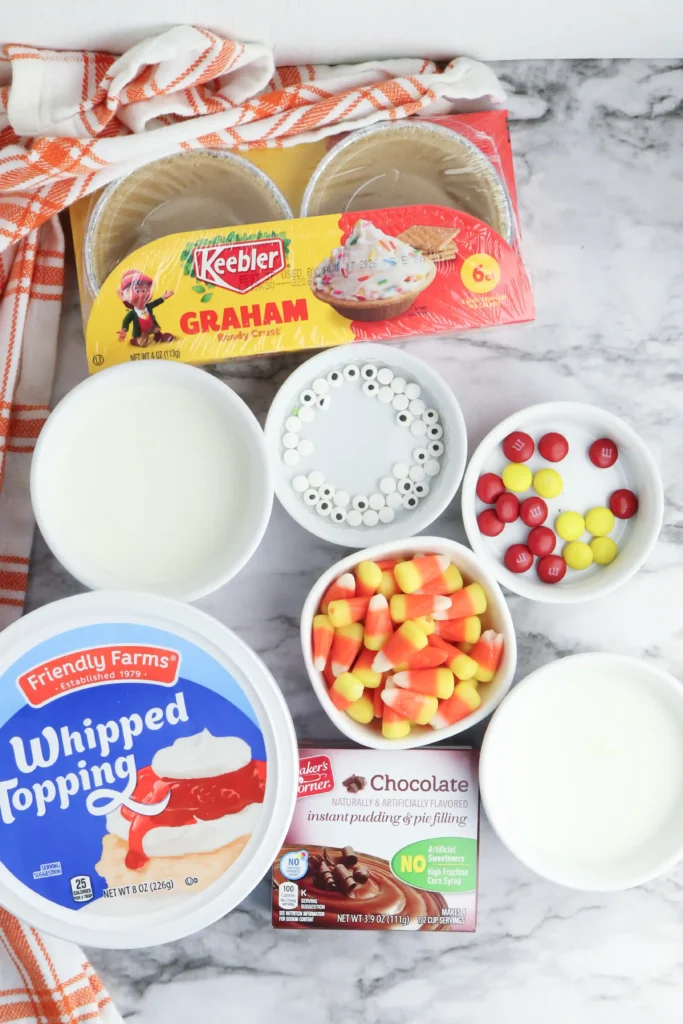 ingredients for the mini turkey pies: Assorted dessert ingredients on a marble counter: graham crackers, whipped topping, candy corn, white nonpareils, and red/yellow candies with an orange striped towel nearby.