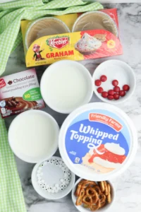 ingredients for the classroom christmas treats: Assorted dessert toppings and snacks on a marble surface with a yellow Keebler Graham Cracker package and green checkered cloth nearby.