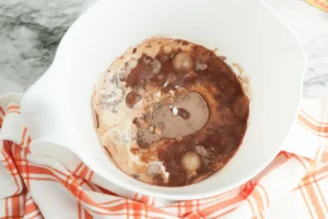 White bowl holding a chocolate dessert with cocoa powder, nuts, and a light cream swirl on a marble countertop with an orange-striped towel nearby.