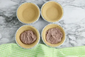 Top-down view of six small pie crusts in aluminum cups on a marble surface, some filled with pudding filling in two of them for these rudolph-themed desserts