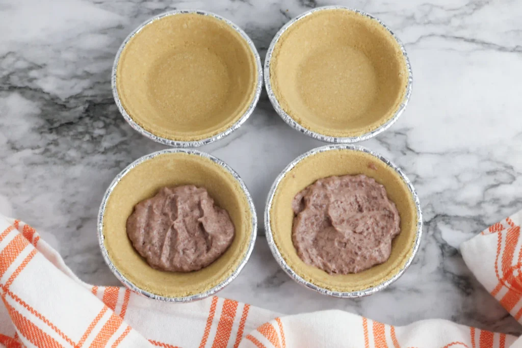 Six foil-lined mini tart or pie crusts arranged in a grid on a marble surface; the bottom two tins contain chocolate pudding batter for these classroom treats for Thanksgiving
