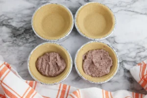 Six foil-lined mini tart or pie crusts arranged in a grid on a marble surface; the bottom two tins contain chocolate pudding batter for these classroom treats