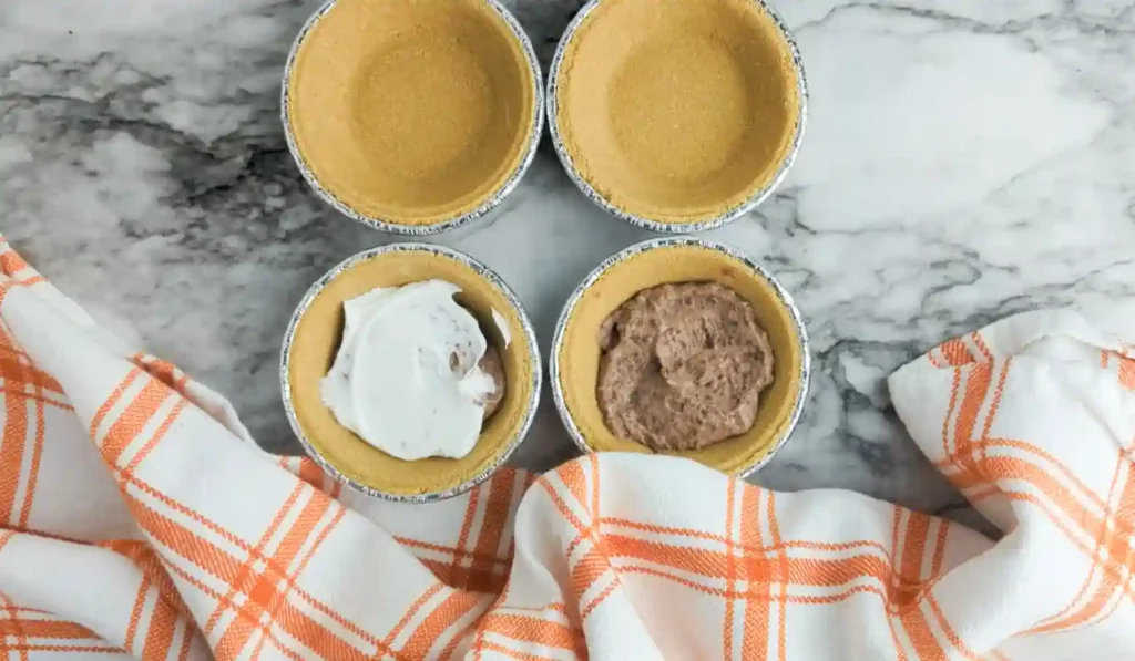 Top‑down view of four mini pie crusts on a marble counter; two are empty, one has white whipped topping and the other brown pudding filling for these turkey-themed desserts