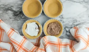 Top‑down view of four mini pie crusts on a marble counter; two are empty, one has white whipped topping and the other brown pudding filling for this turkey-themed dessert