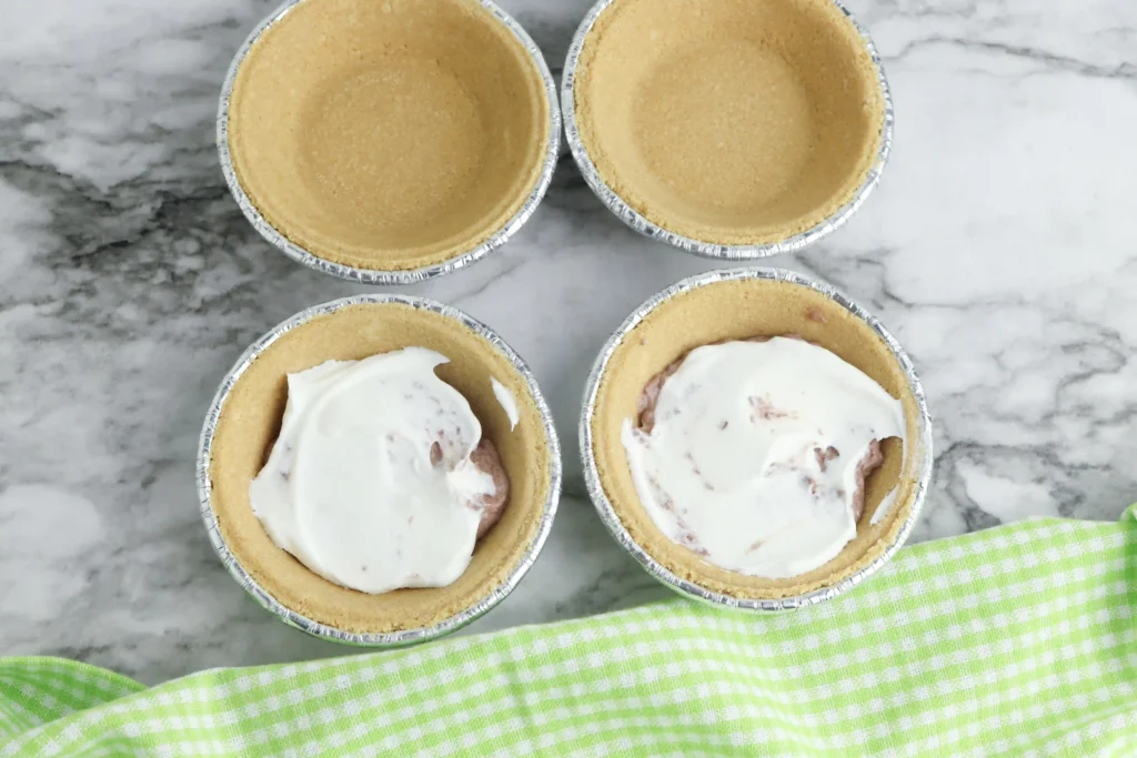 Mini Rudolph Pies - Cultivating Brilliant Minds Top-down view of mini pie crusts in aluminum tins on a marble surface; two of them having chocolate pudding and whipped topping in them