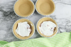 Top-down view of mini pie crusts in aluminum tins on a marble surface; two of them having chocolate pudding and whipped topping in them