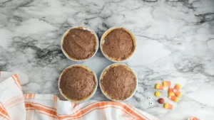 Four mini chocolate pies in aluminum pans arranged in a 2x2 grid on a marble countertop, with an orange-striped towel nearby and candy on the right side.
