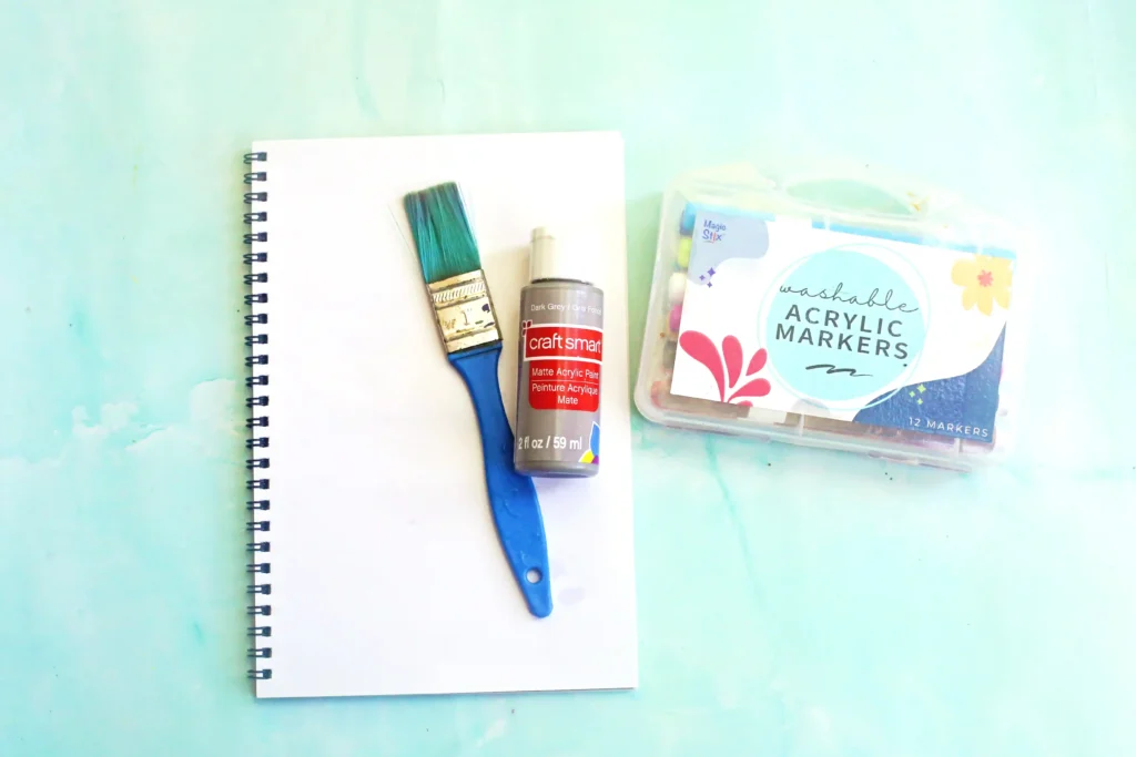 supplies for this animal craft for kids: Blue-handled paintbrush, bottle of matte acrylic paint, and a pack of washable markers on a notebook sheet on a pale blue background.