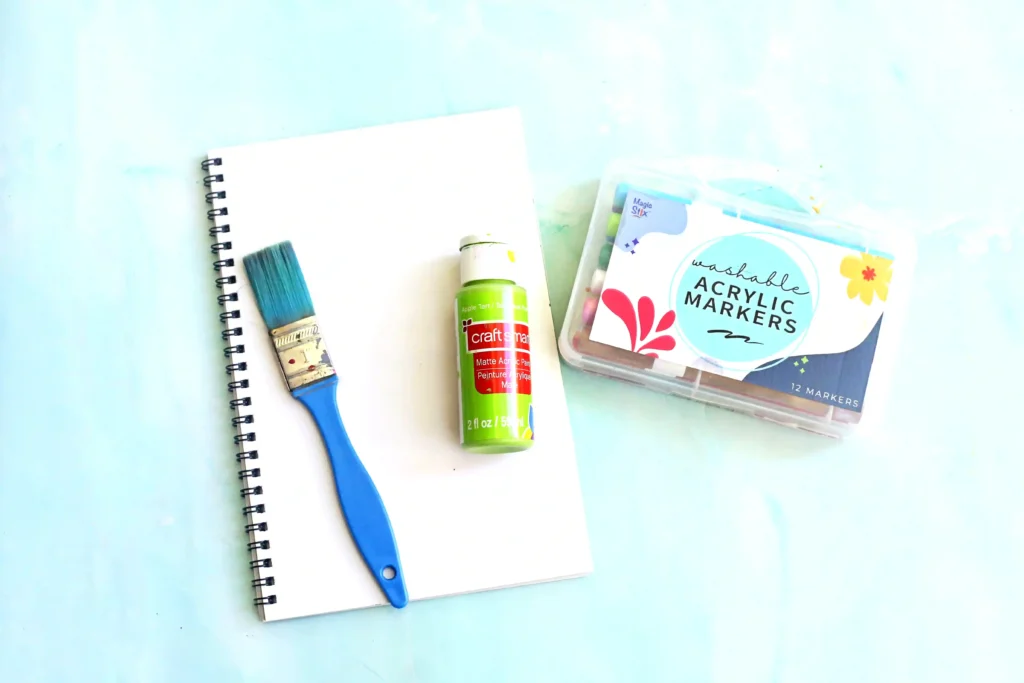 Letter L Leaf Handprint Craft - Cultivating Brilliant Minds supplies for this handprint art: Flat lay of a spiral notebook, blue paintbrush, green acrylic paint bottle, and a pack of washable acrylic markers on a light blue surface.