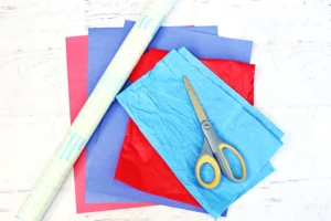 supplies for the lego brick suncatcher: colorful sheets of paper (red, blue, pink) with scissors and a roll of patterned wrapping paper on a light surface.
