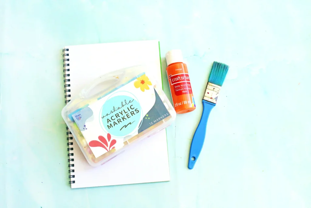 Letter M Handprint Craft - Cultivating Brilliant Minds supplies for this monster craft for kids: Orange acrylic paint bottle with white cap beside a blue paint brush on a pale background and a spiral notebook nearby.
