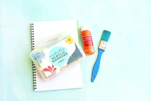 supplies for this monster craft for kids: Orange acrylic paint bottle with white cap beside a blue paint brush on a pale background and a spiral notebook nearby.