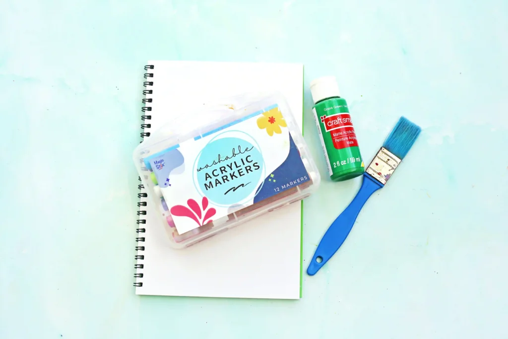 supplies for this letter N handprint craft: Open spiral notebook with a pack of washable acrylic markers and a green Craft Smart paint bottle beside a blue brush on a light blue background, ready for art.