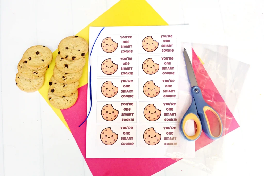 Cookies stacked on a yellow board beside a sheet of'You're one smart cookie' tags with a cookie image and scissors nearby.