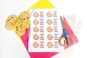 Cookies stacked on a yellow board beside a sheet of 'You're one smart cookie' tags with a cookie image and scissors nearby.