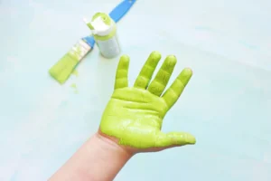 Green-painted hand raised in the foreground with a blue and green paint bottle and brush blurred in the background for this alphabet craft for kids