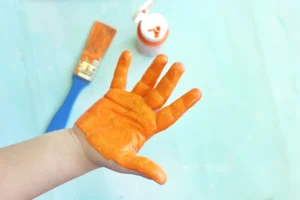 Hand coated in bright orange paint, held up with a paintbrush and orange paint bottle visible in the background on a light blue surface.