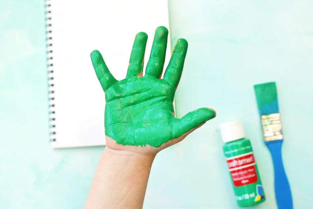 Hand raised with bright green paint covering palm and fingers, notebook and paint bottles visible in the background while crafting.
