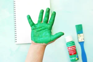 Hand raised with bright green paint covering palm and fingers, notebook and paint bottles visible in the background while crafting.