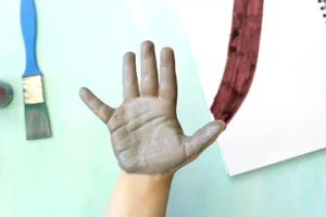 Child's hand covered in gray paint during painting activity, with a paintbrush nearby on a light surface for this handprint art