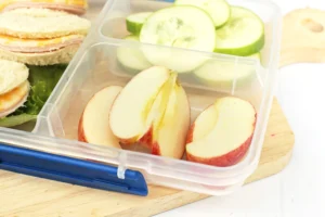 Lunchbox with sliced apples, cucumber rounds, and a sandwich with lettuce visible inside a clear container