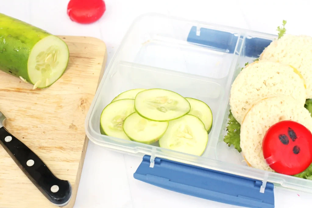 Very Hungry Caterpillar Lunch Box Idea - Cultivating Brilliant Minds Lunchbox with cucumber slices on a white surface, a cucumber half on a cutting board, and part of a sandwich with tomato edges in view.