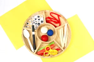 supplies for the Toy Story Forky Playdough Tray- Round wooden craft organizer filled with googly eyes, scissors, markers, and colorful clay on a bright yellow background.