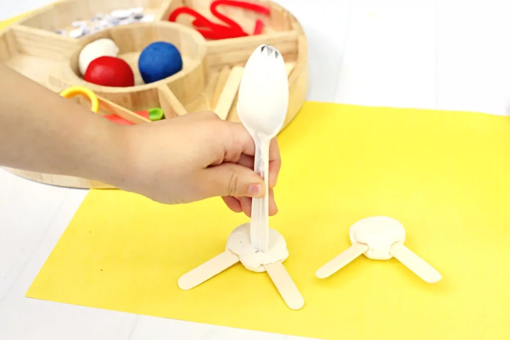 Toy Story Forky Playdough Tray - Cultivating Brilliant Minds Child's small hand holds a white plastic spoon, assembling clay shapes on wooden craft sticks on a bright yellow surface.