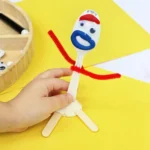 Close-up of a hand holding a DIY toy story forky made from a plastic spoon, with a blue mouth, red eyebrows, and red pipe cleaner arms on a yellow surface.
