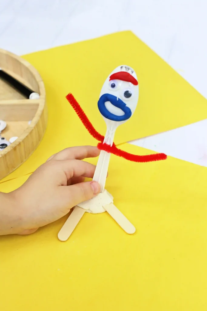 Toy Story Forky Playdough Tray - Cultivating Brilliant Minds Close-up of a hand holding a DIY toy story forky made from a plastic spoon, with a blue mouth, red eyebrows, and red pipe cleaner arms on a yellow surface.