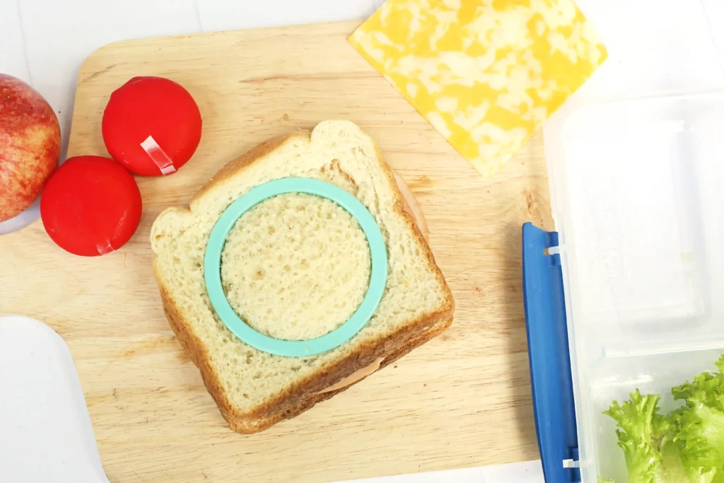 Very Hungry Caterpillar Lunch Box Idea - Cultivating Brilliant Minds Bread slice on a wooden board with a turquoise circular food mold, two red tomatoes, a yellow cheese block, and a clear container nearby for these kid lunchbox ideas