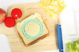 Bread slice on a wooden board with a turquoise circular food mold, two red tomatoes, a yellow cheese block, and a clear container nearby.