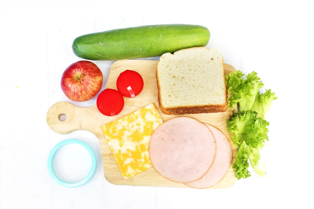 Very Hungry Caterpillar Lunch Box Idea - Cultivating Brilliant Minds ingredients for the very hungry caterpillar lunch box idea: Top-down view of a cutting board with cucumber, apple, red tomatoes, sliced bread, ham, cheese, and lettuce.