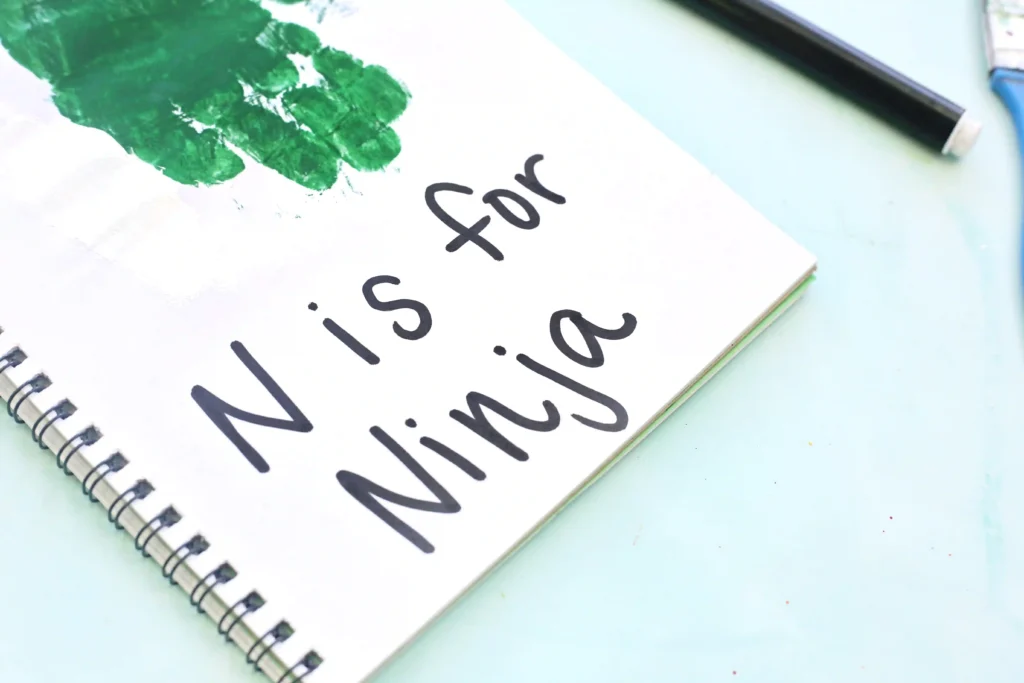 Close-up of a spiral notebook on a pale blue surface, with green paint on the page and large black handwritten words visible.